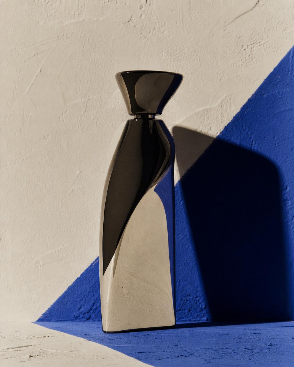 A polished sculptural object hit with hard light against a cream wall with a slash of deep indigo