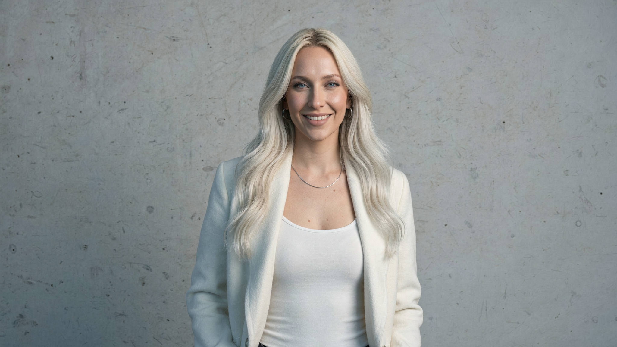 Taryn smiling in an open cream wool coat against a concrete wall in cool daylight.